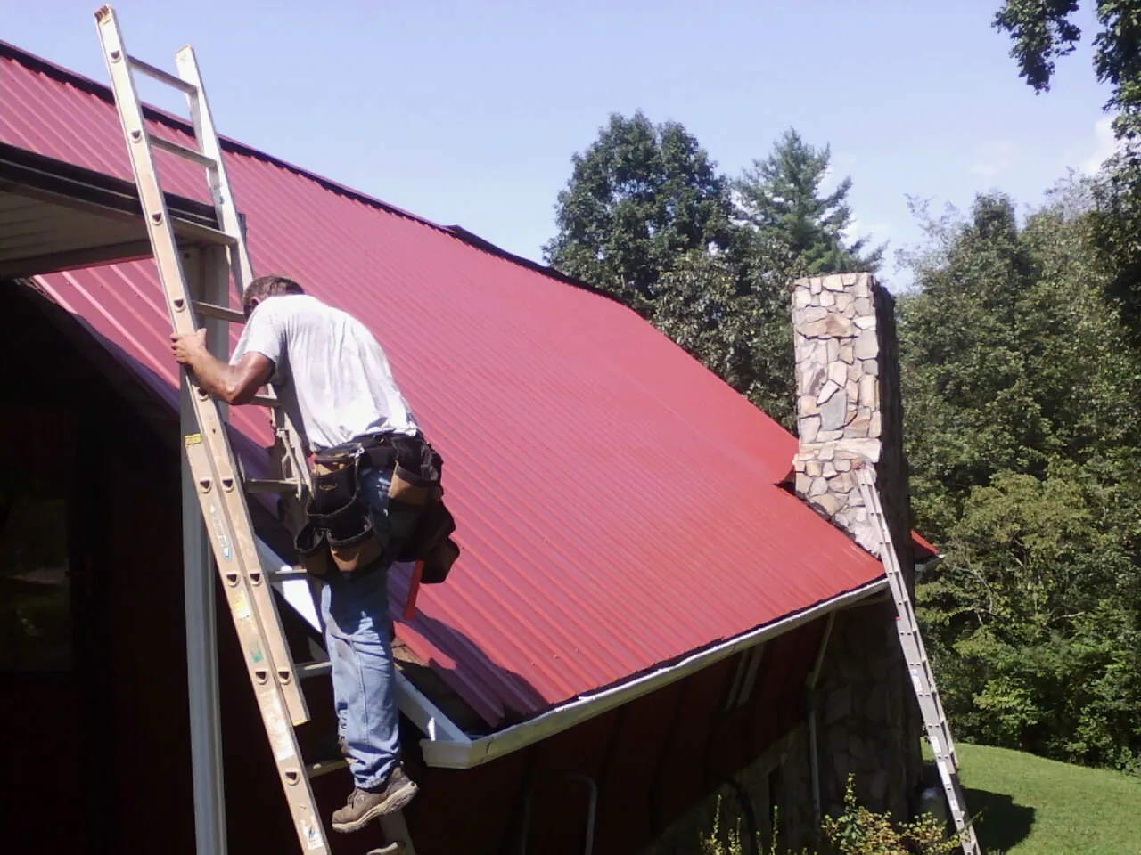 Roofer on a ladder beside a red metal roof and stone chimney — Soffit & Fascia Repair by Roofing Brothers in Bosler